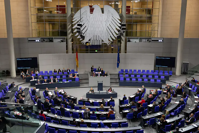 Das Bild zeigt, wie eine Debatte im Bundestag stattfindet. Ein Abgeordneter steht vorne und hält eine Rede. Die anderen Abgeordneten sitzen auf ihren Plätzen in einem Halbkreis um das Rednerpult.