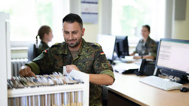Die Grafik zeigt einen Soldaten der Bundeswehr in einem Büro bei Verwaltungs- und Organisationsaufgaben. Er sitzt an einem Schreibtisch und sortiert Akten in einem Karteischrank. Im Hintergrund arbeiten weitere Soldatinnen und Soldaten an Computern. Die Szene veranschaulicht typische Tätigkeiten im Stabsdienst, bei denen Büroorganisation, Dokumentenverwaltung und administrative Aufgaben im Mittelpunkt stehen.