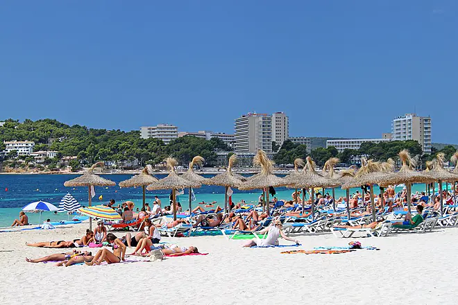 Ein Bild zeigt einen belebten Strand auf Mallorca. Im Hintergrund stehen einige große Hotelbauten.