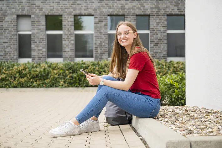 Duale Studentin sitzt im Außenbereich des Büros mit Handy in der Hand