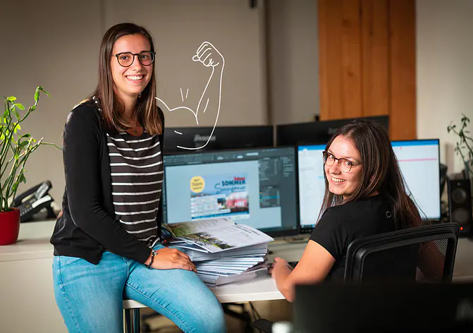 Zwei Frauen in einem Büro, eine sitzt am Schreibtisch vor zwei Bildschirmen, die andere lehnt sich mit einem Stapel Papier auf dem Schoß an den Tisch, an der Wand ist eine gezeichnete Muskelflex-Grafik zu sehen.