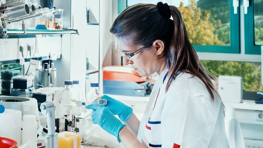 Eine Laborantin mit Schutzbrille und blauen Handschuhen arbeitet konzentriert mit einer Pipette an einer Arbeitsstation in einem hellen, modern ausgestatteten Labor