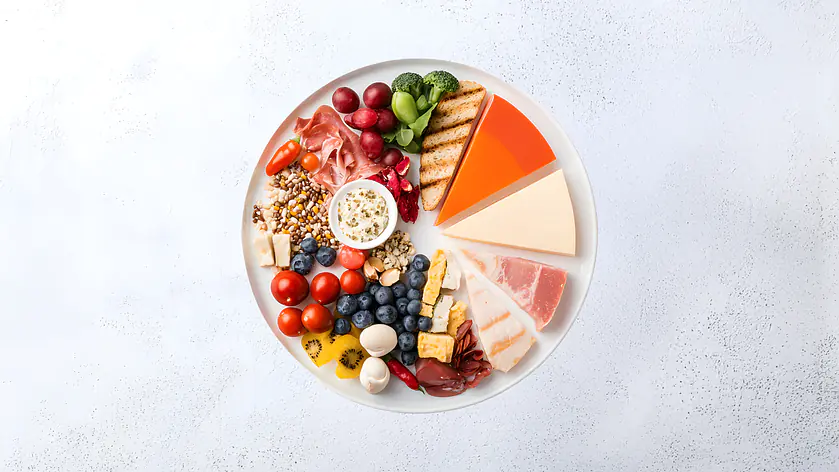 Draufsicht auf einen Teller mit vielfältigen Lebensmitteln wie Käse, Obst, Gemüse, Nüssen, Getreide und Aufschnitt, angeordnet in Segmenten