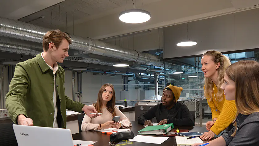 Projektarbeit auf Augenhöhe: Eine studentische Lerngruppe im lebendigen Austausch an einem großen Arbeitstisch mit Laptops und Unterlagen