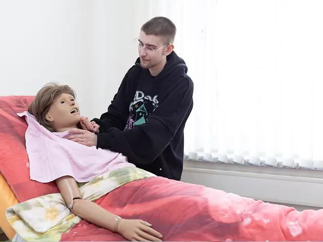 Der Mann steht neben einer Übungspuppe auf einem Bett. Die Puppe ist mit Tüchern und einer Decke bedeckt. Im Hintergrund ist ein helles Fenster.