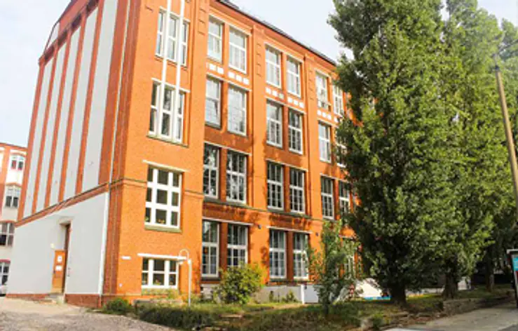 Ein mehrstöckiges Backsteingebäude mit vielen großen Fenstern. Rechts davor stehen hohe grüne Bäume. Vor dem Gebäude gibt es einen kleinen Gartenbereich und einen gepflasterten Weg.