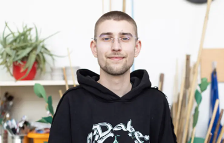 Ein Mann mit kurzer Frisur und Brille steht vor Regalen mit Pflanzen, Pinseln und Holzstäben. Er trägt einen schwarzen Hoodie.