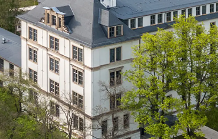 Ein helles mehrstöckiges Gebäude mit dunklem Dach. Viele hohe Fenster sind sichtbar. Im Vordergrund und rechts stehen grüne Bäume. Das Foto ist von oben aufgenommen.