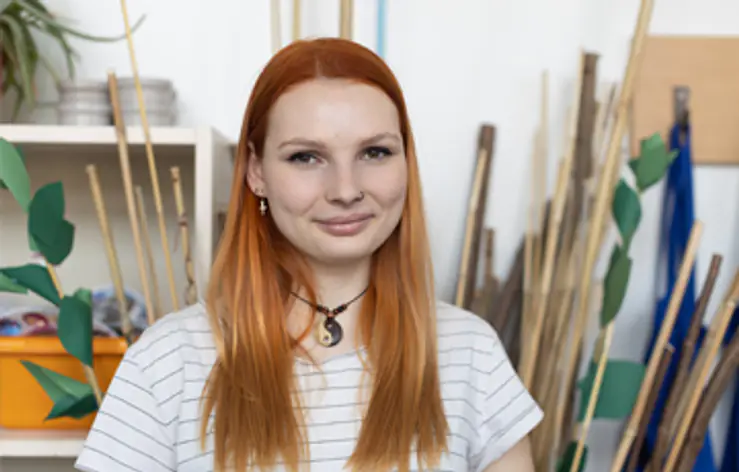 Eine junge Frau mit langen roten Haaren steht vor einer hellen Wand. Sie trägt ein weißes, gestreiftes T-Shirt und eine Halskette. Im Hintergrund stehen viele Holzstäbe und einige grüne Blattformen.