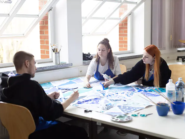 Drei Personen sitzen an einem großen Tisch. Auf dem Tisch liegt ein großes Blatt mit blauen Mustern. Die Personen malen oder arbeiten daran. Der Raum ist hell und hat große Dachfenster.