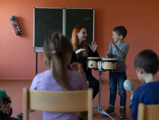 Eine Frau und ein Junge sitzen an zwei Trommeln. Sie lächeln sich an und heben die Hände. Im Vordergrund sitzen mehrere Kinder. Im Hintergrund steht eine Tafel vor einer roten Wand.