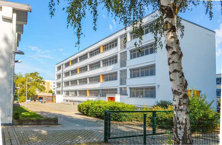 Ein großes weißes Gebäude mit mehreren Etagen und vielen langen Fenstern. Vor dem Gebäude stehen Büsche, ein grüner Zaun und eine Birke. Links verläuft ein gepflasterter Weg. Der Himmel ist blau.