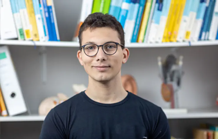 Ein junger Mann mit kurzer dunkler Frisur und Brille steht vor einem Bücherregal. Er trägt ein dunkles T-Shirt. Im Regal stehen viele Ordner und Bücher.