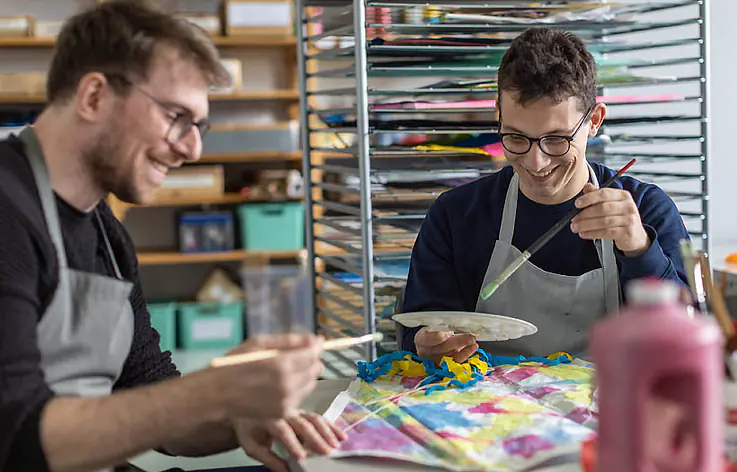 Zwei Männer sitzen an einem Tisch und malen. Sie tragen Schürzen und halten Pinsel. Auf dem Tisch liegen bunte Farben und ein bemaltes Blatt.