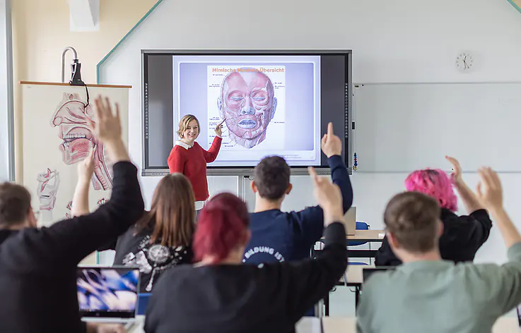 Eine Lehrerin steht vor einer großen digitalen Tafel. Auf der Tafel ist eine medizinische Zeichnung eines Gesichts zu sehen. Mehrere Personen sitzen im Raum und heben die Hände.