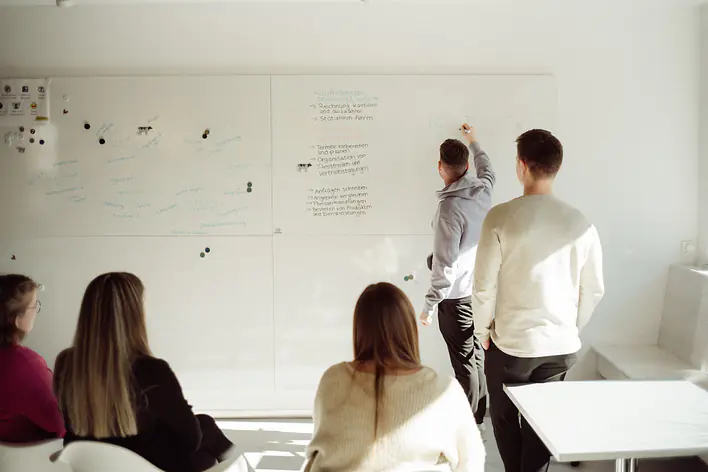 Workshop am Whiteboard Eine Gruppe verfolgt eine Präsentation oder Diskussion vor einem großen Whiteboard. Zwei Personen stehen vorne und ergänzen bzw. erläutern die dort notierten Inhalte.