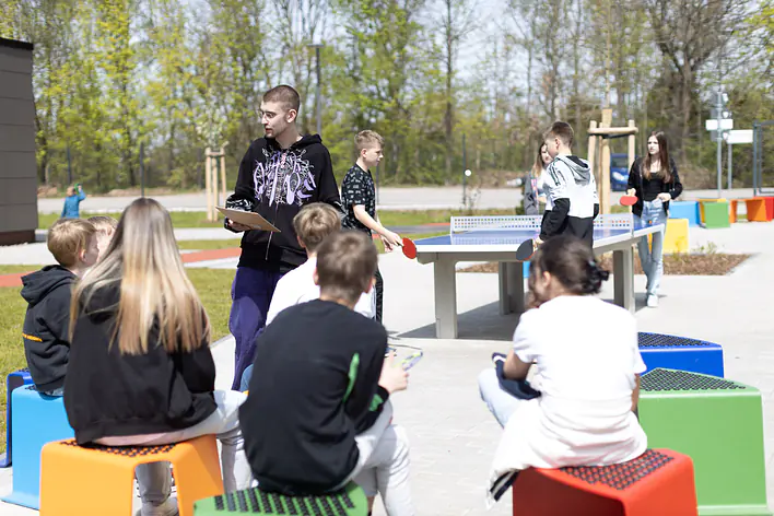 Ein Mann steht auf einem Schulhof. Vor ihm sitzt eine Gruppe von Kindern. Im Hintergrund spielen Kinder Tischtennis.
